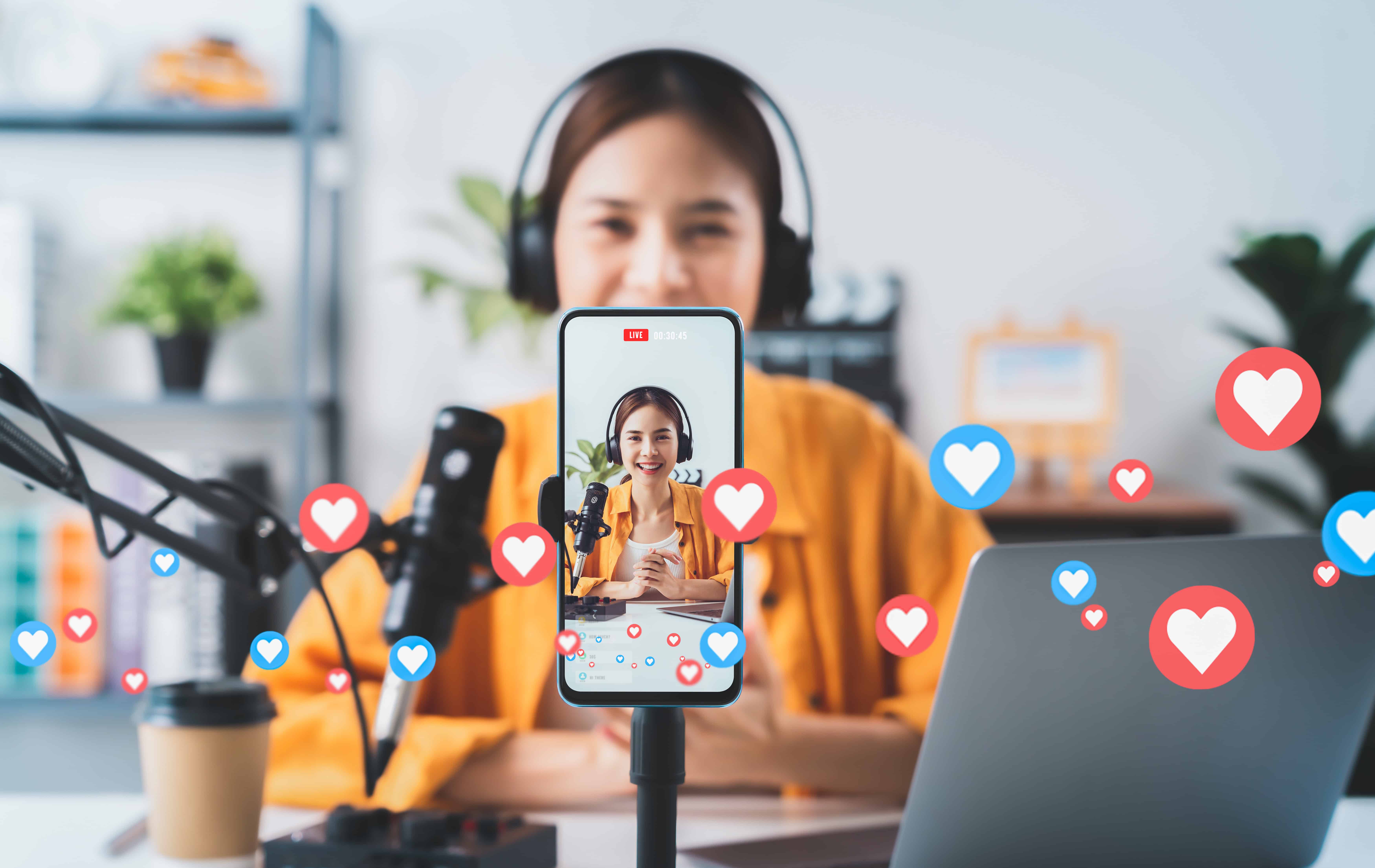 A young woman live-streaming on her phone with social media engagement icons, like hearts and thumbs-up, surrounding her.