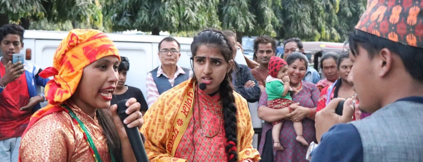 A group of young people in Nepal, including two women with microphones and one man in traditional attire, lead an anti-trafficking awareness campaign, surrounded by an engaged community audience.