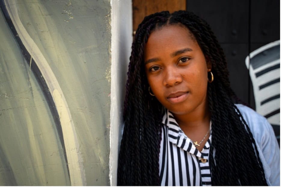 A girl with braids stands in front a green painted wall. She is wearing a striped shirt.