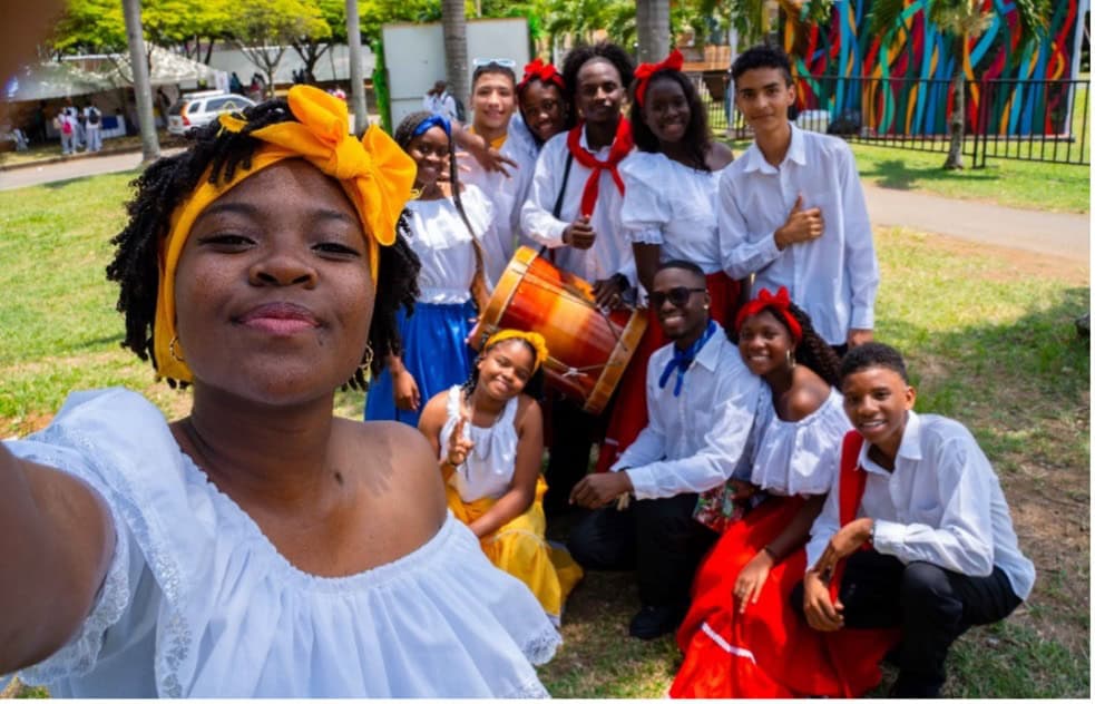 A group of people gather around a drum. They are wearing colorful, traditional Colombian dress and are standing in a park.