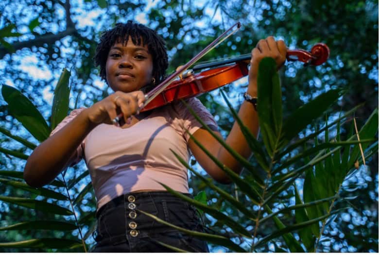 A girl in a beige shirt and skirt with buttons with twisted hair plays the violin in a jungle.