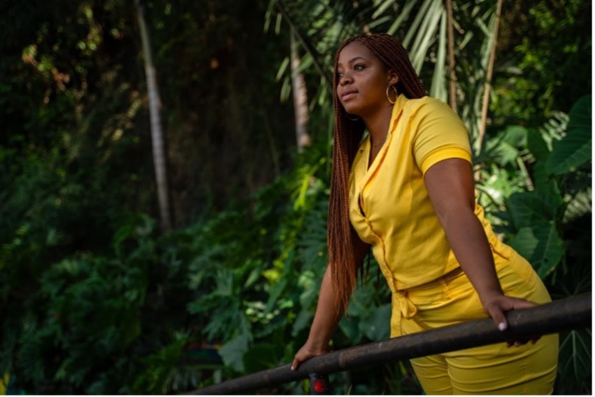 A woman with long brown hair stands in a yellow dress leaning against a railing. A jungle is behind her.