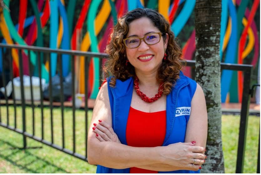 A women with curly brown hair stands in front of a fence and a wall mural painted with colorful lines. She is wearing brown glasses, and has a red necklace made of beads and a blue vest with the Plan International logo.