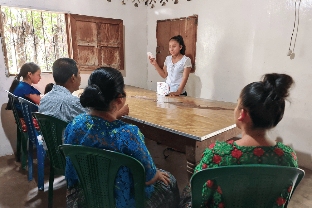 A girl stands in front of a long, brown table. Four other people look at her from the other side. She is holding menstrual pads.