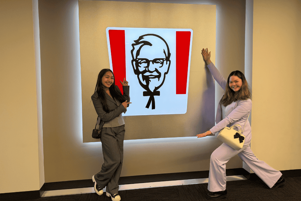 Two young women in suits stand in front of a wall that has an illuminated version of the KFC logo.