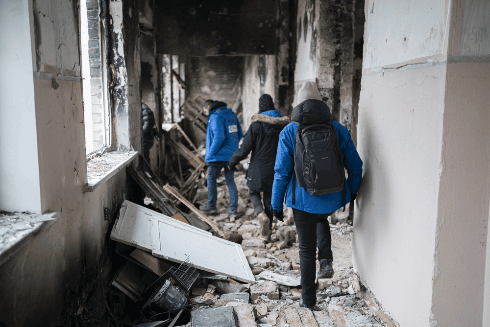 Three people walk down a crumbling, fire-damaged hallway in a school in Ukraine. All three are facing away from the camera.