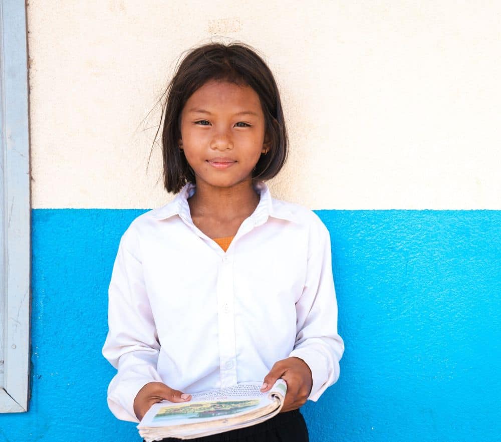 A girl with short black hair wearing a white blouse stands in front of a blue and white wall, holding a book and smiling.