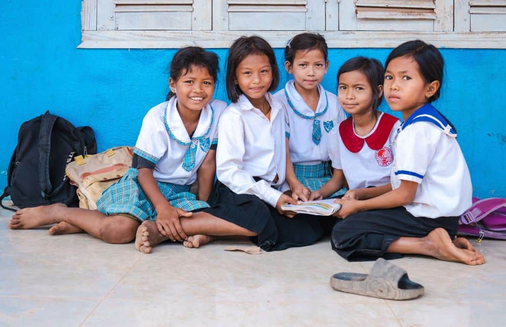 Five girls sit in front of their school building, holding a textbook and looking at the camera.