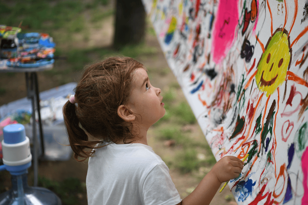 A girl in a white t-shirt sits in front of a mural and paints swirls and hearts.