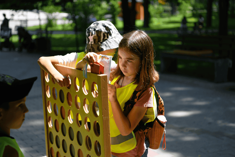 A girl wearing a bright yellow vest stands and looks determined at a game of Connect Four. A boy wearing a camo hat is next to her, looking away from the camera. Another boy can be seen in the bottom left corner of the foreground.