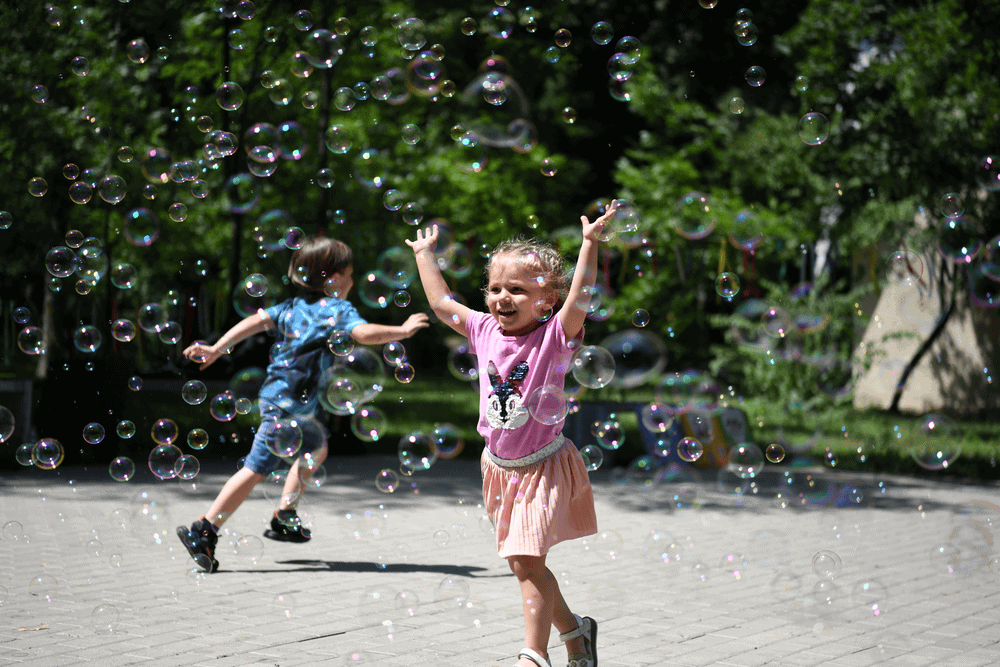 In the foreground, girl wearing pink jumps through a cloud of bubbles. In the background, a boy wearing blue runs in the opposite direction, facing away from the camera.