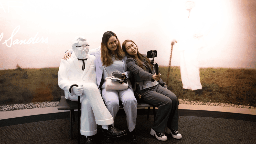 Pear and Shirlyn sit with the Colonel Sanders statue holding a phone on a selfie stick.