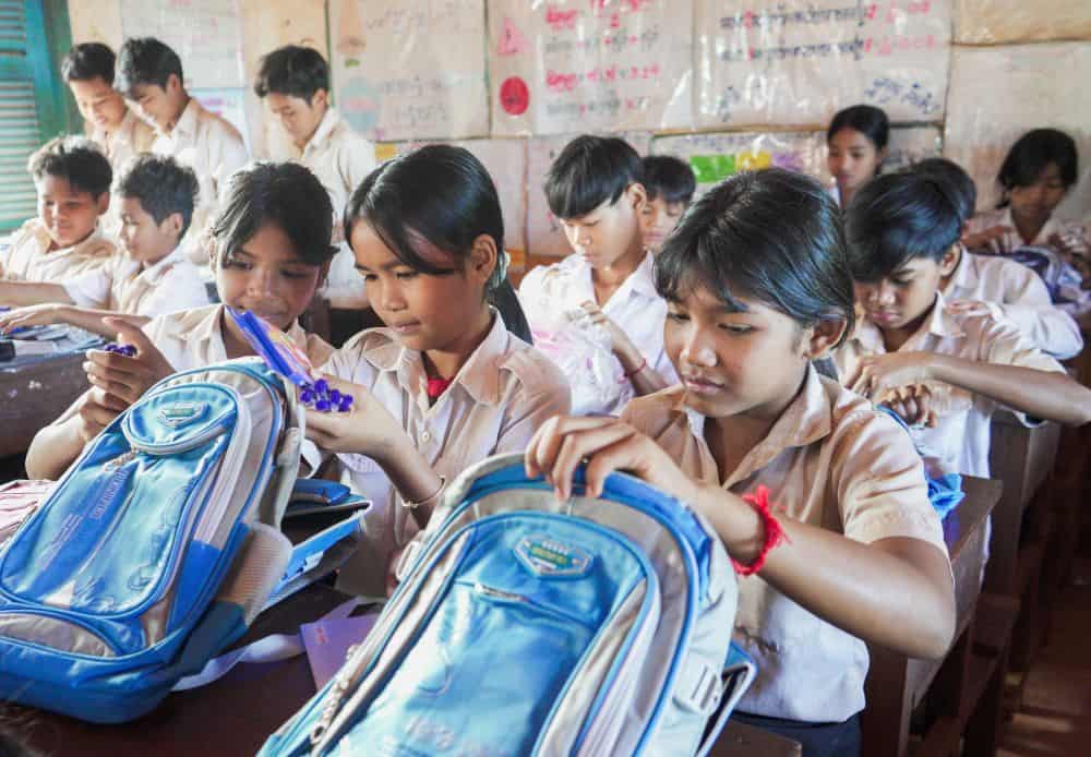 Students in a classroom in Cambodia sit at their desks and look at the school supplies in their new blue backpacks.