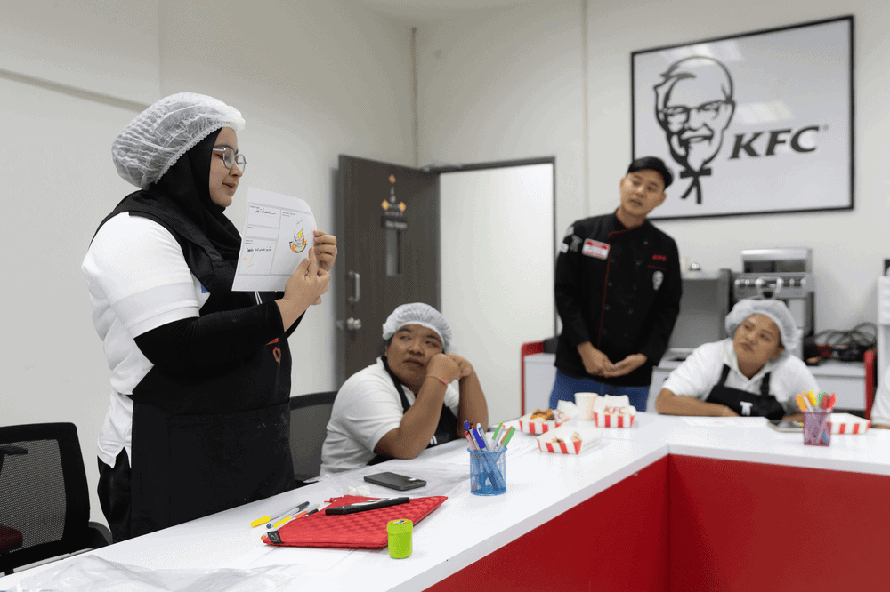 Aliya holds a piece of paper to present a product concept idea to a table of KFC employees.