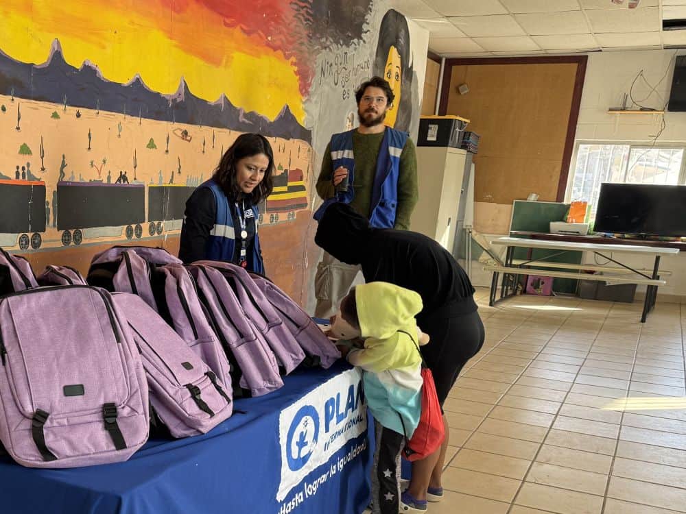 Purple backpacks are arranged on a blue table with a Plan banner. A parent and child sign in to receive their backpack.