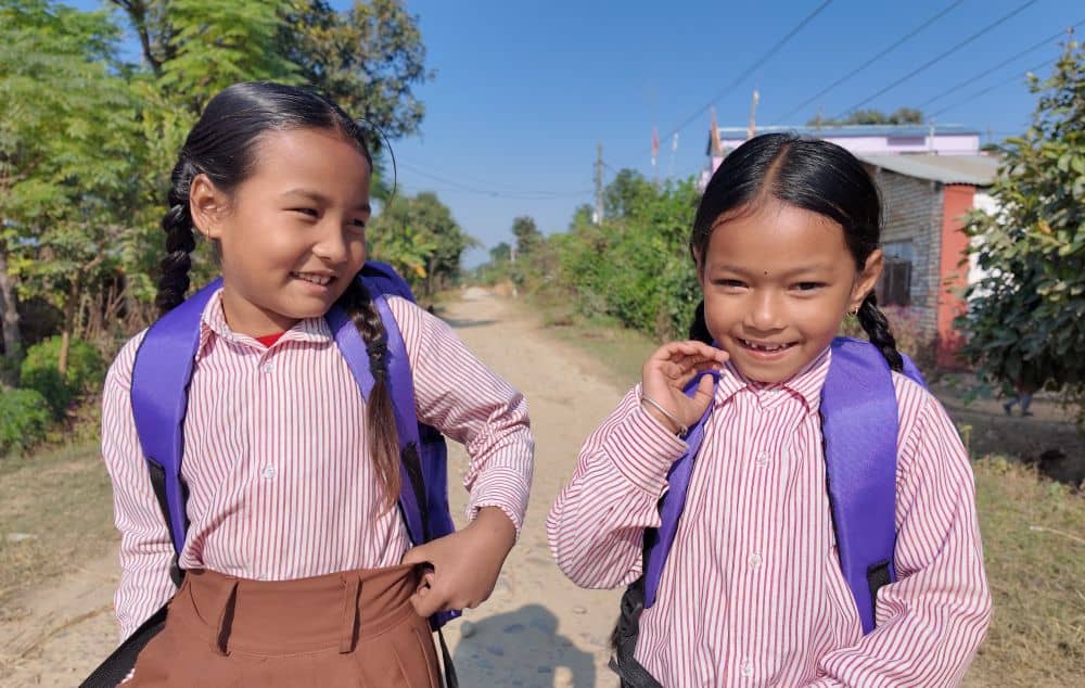 Two girls with dark hair stand in a dirt road and smile. They are wearing striped pink and white blouses and purple backpacks.