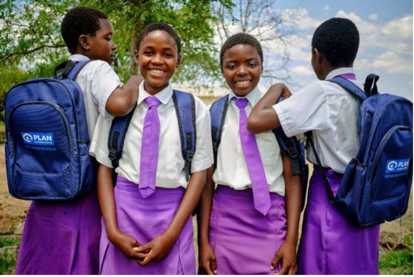 Four girls in purple skirts with purple ties stand next two each other. Two face front and two face away, wearing backpacks with Plan's logo.