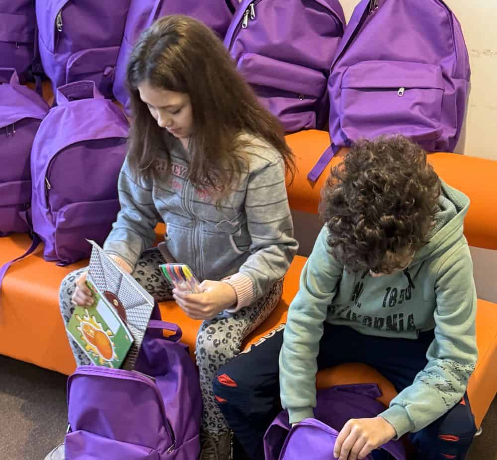 A girl and boy in Ukraine sit on an orange bench next to purple backpacks, holding school supplies. 