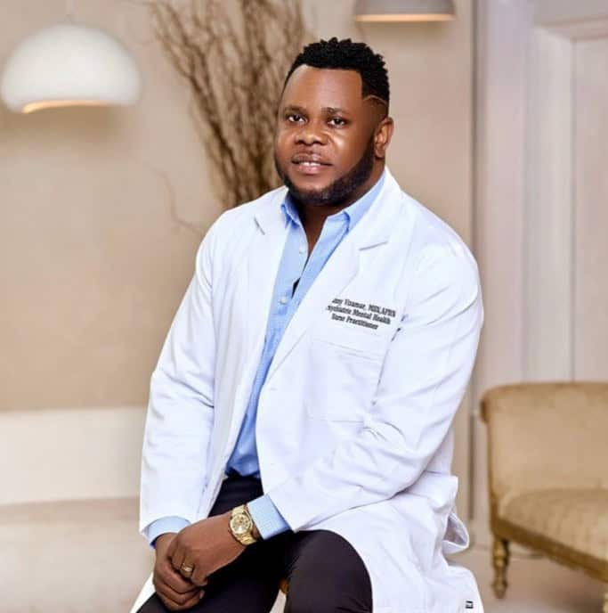 A man sits on a stool and looks at the camera. He is wearing a white medical coat.