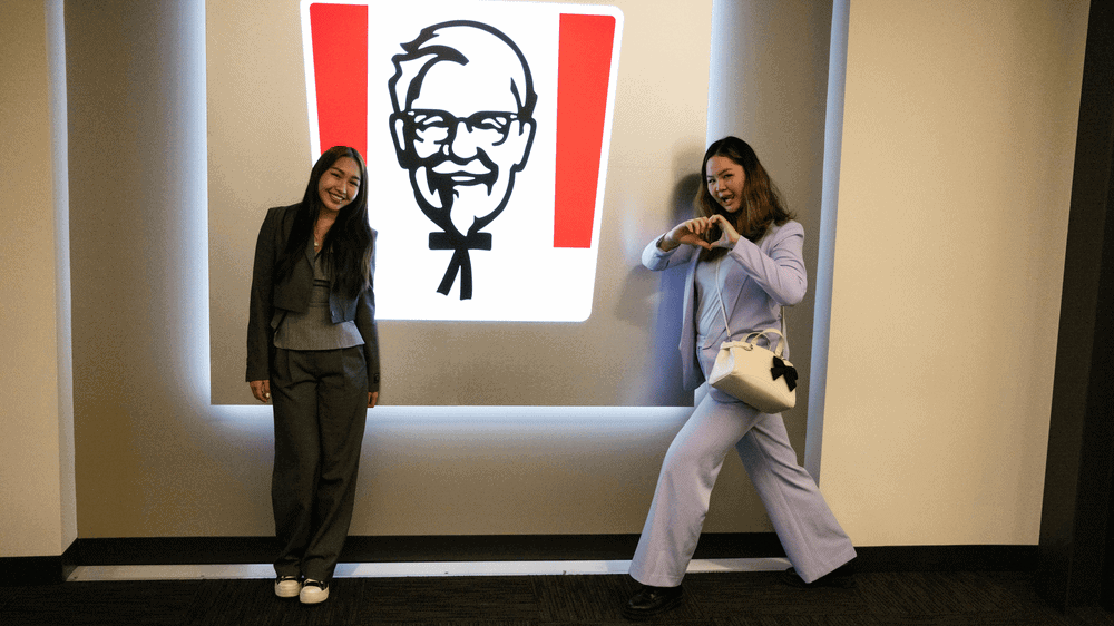 Two young women stand in front of a giant sign with the face of KFC's Colonel Sanders.