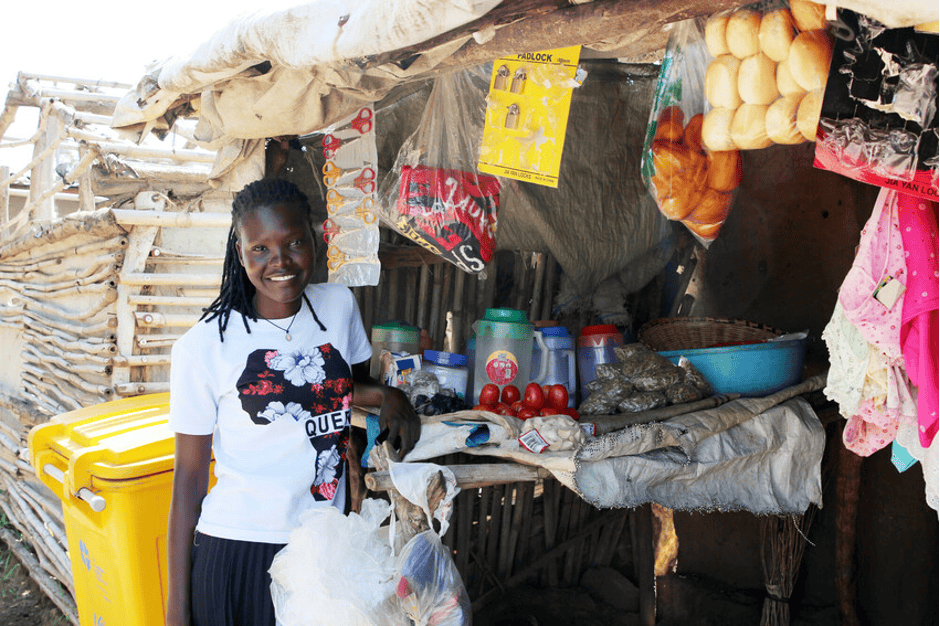 Ruth stands in front of her small storefront. There are bags hanging from the roof with fruit, fabric and clothes, and a table she is leaning on with other goods for sale.
