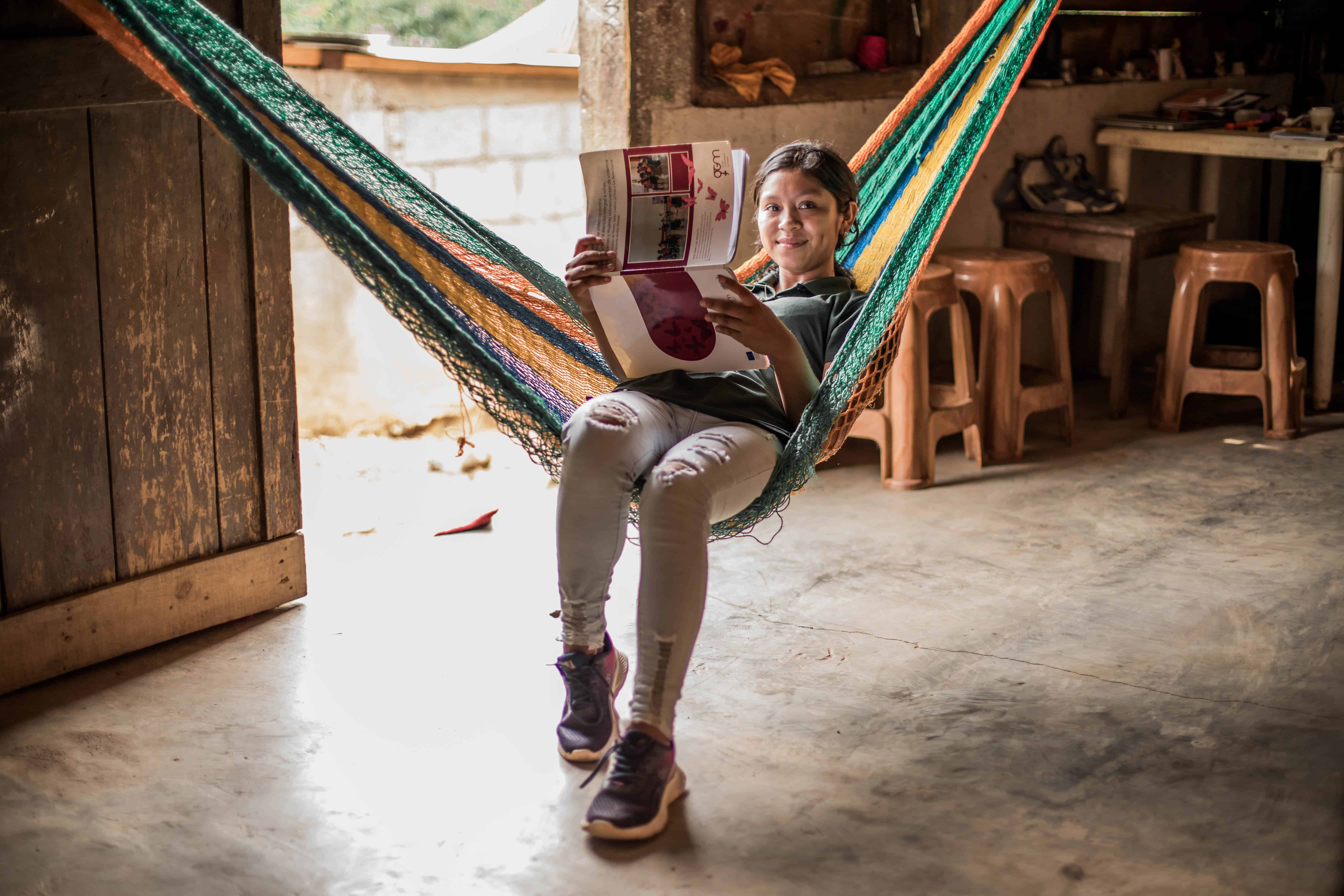A young girl in Guatemala sits in a colorful hammock, smiling as she reads a booklet. The leadership school helps girls like her develop advocacy and leadership skills.