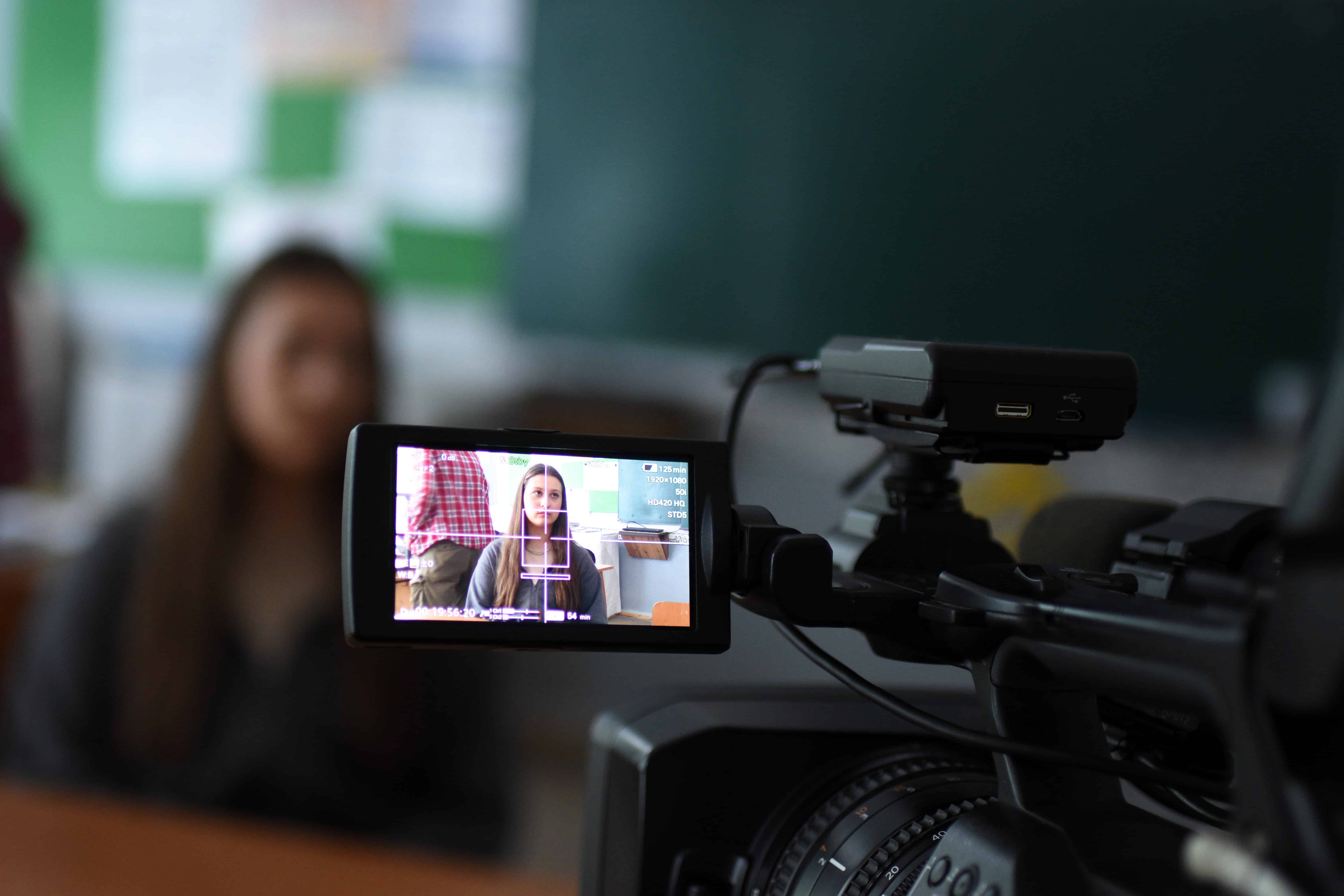A video camera captures a young girl, Maria, being interviewed in a classroom setting. The blurred background shows a chalkboard and classroom environment. Maria, a 15-year-old Ukrainian refugee, shares her experience of adjusting to life in Moldova after being displaced by conflict.