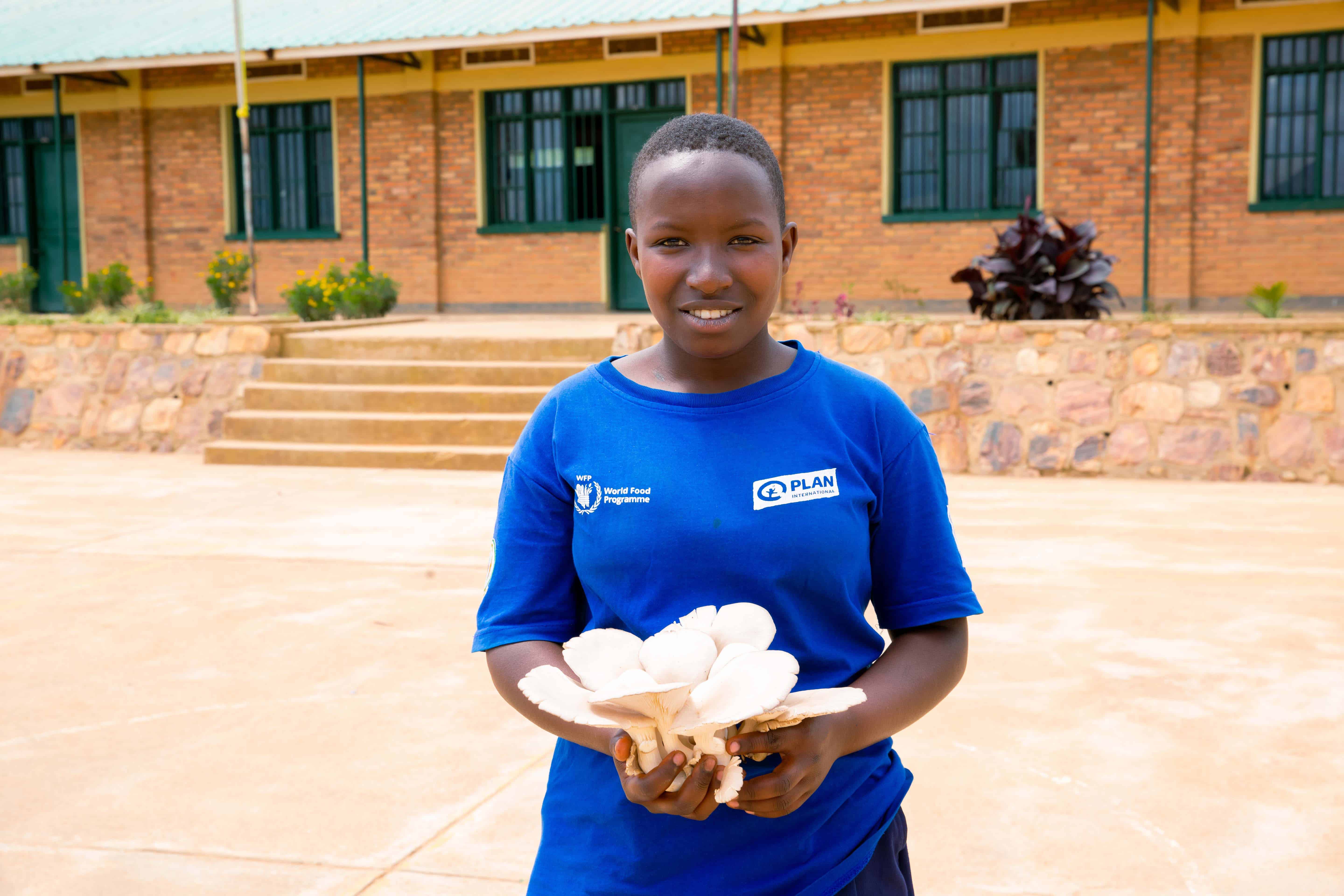A 16-year-old girl named Alice, wearing a blue Plan International shirt, stands outside her school in Rwanda, smiling while holding a handful of freshly harvested mushrooms. She leads a student nutrition club that grows food to combat malnutrition in local communities.