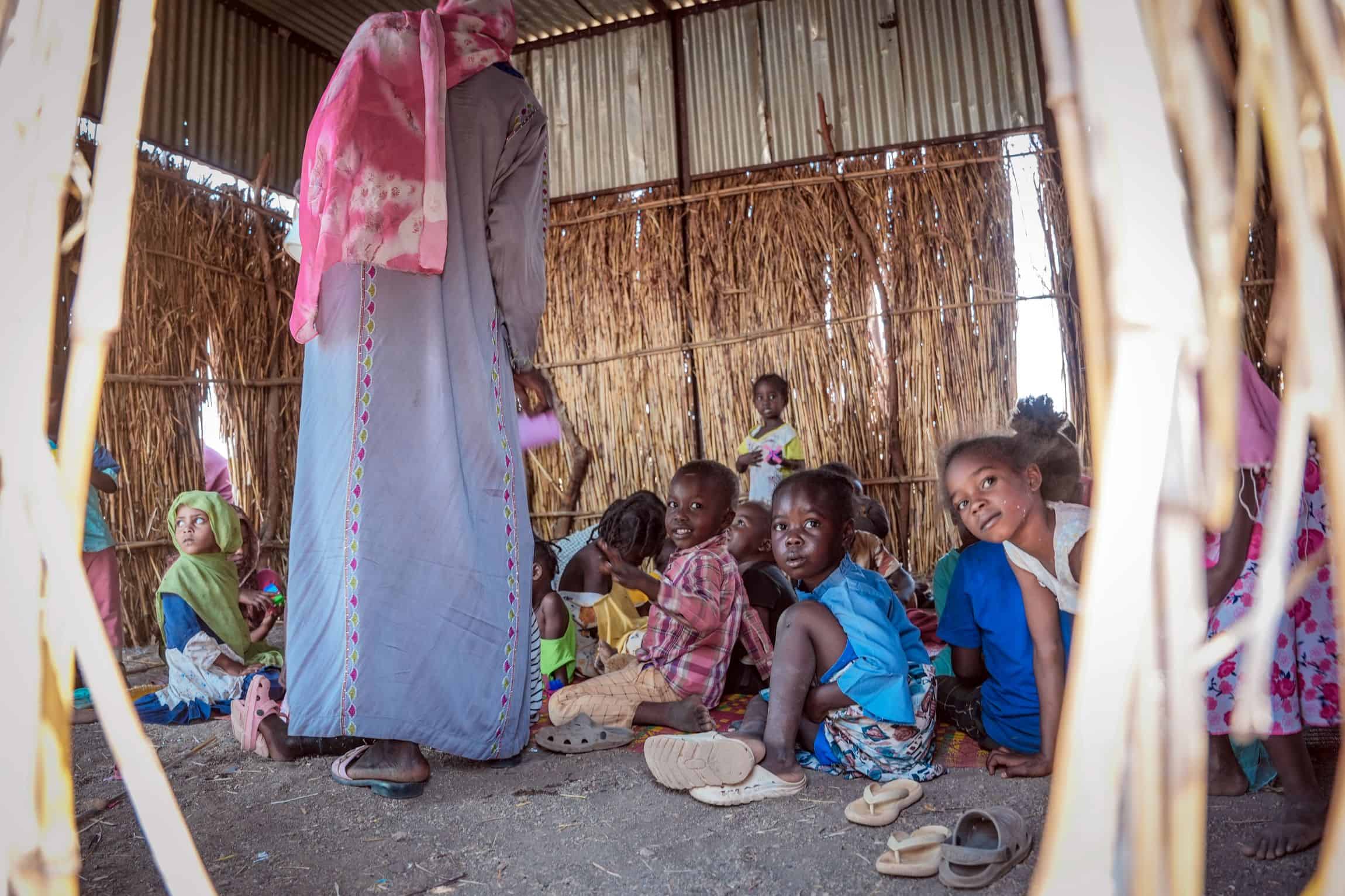 A group of young children sit on the floor of a thatch-and-tin shelter, looking toward the camera. They are inside a child-friendly space at an IDP camp in Gadarif, Sudan. A woman wearing a long dress and headscarf stands nearby.