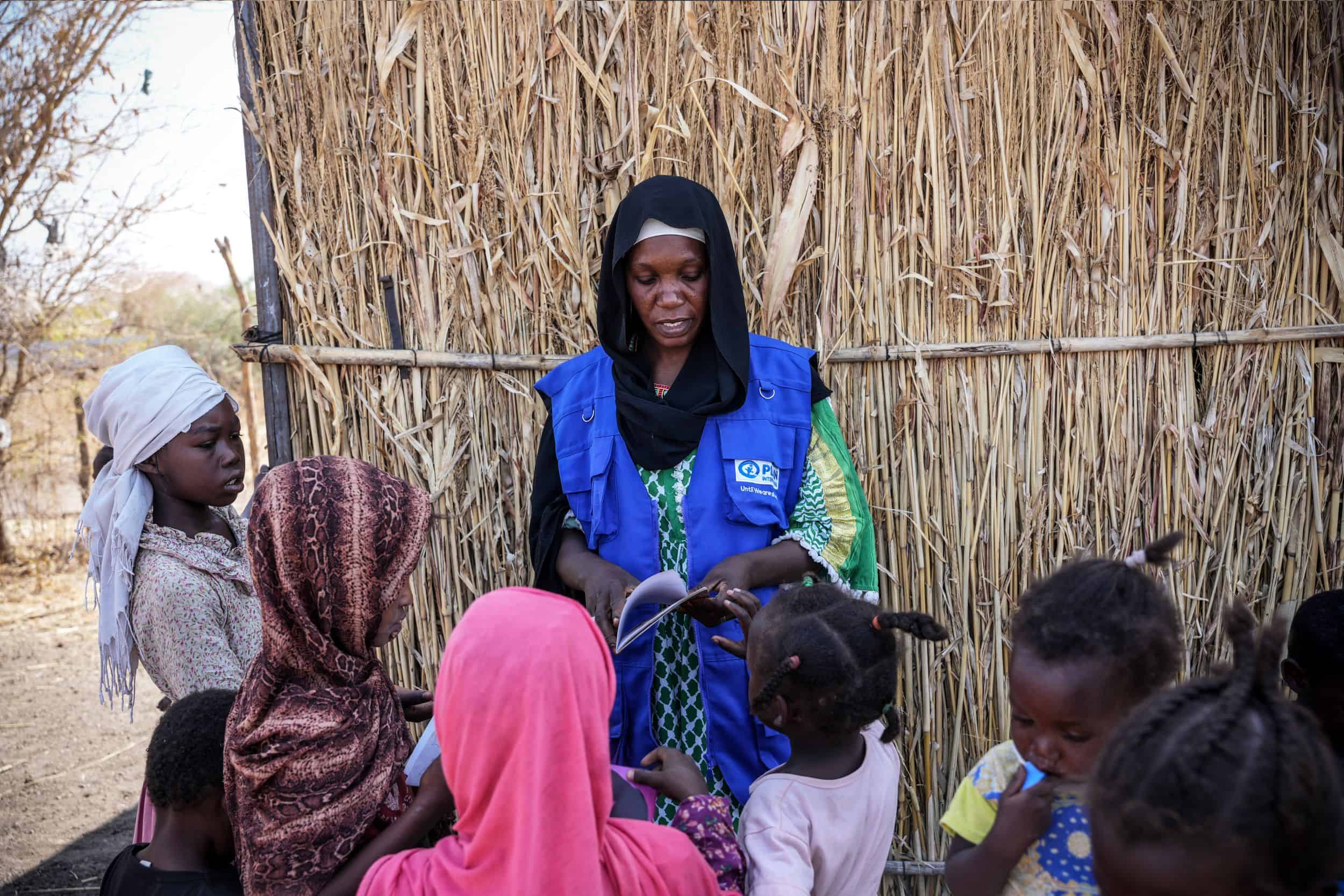 Nasra Abdalla, a child-friendly space facilitator, reads a storybook to children at an IDP camp in Gadarif state, Sudan. She wears a black headscarf and a blue Plan International vest. Several children sit and listen closely.