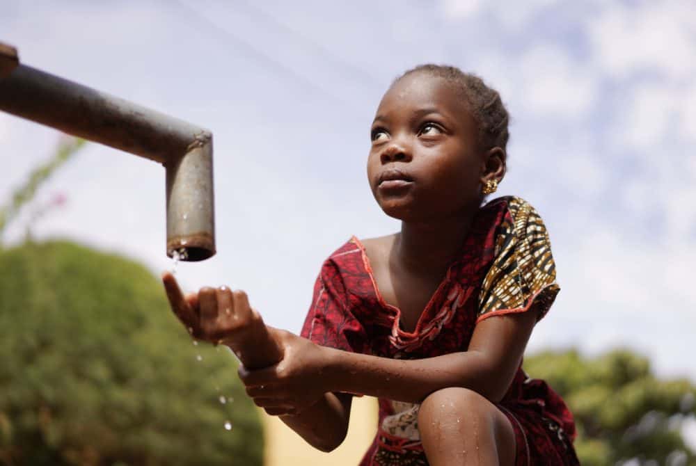 Young girl in patterned dress collecting water from a nearly empty tap outdoors.