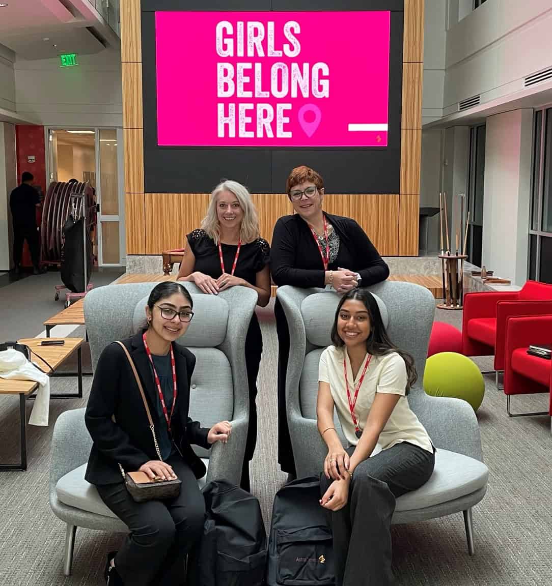 Soha and another Youth Advisory Board member sit with two AstraZeneca employees during Plan’s Girls Belong Here Takeover. A pink screen behind them reads “Girls Belong Here.” All four women are smiling in a modern office space.
