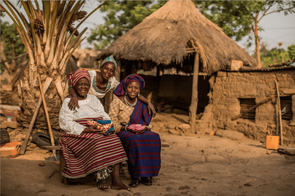 Grandmother, mother and granddaughter in traditional dress sit together in front of a thatched home in Togo.