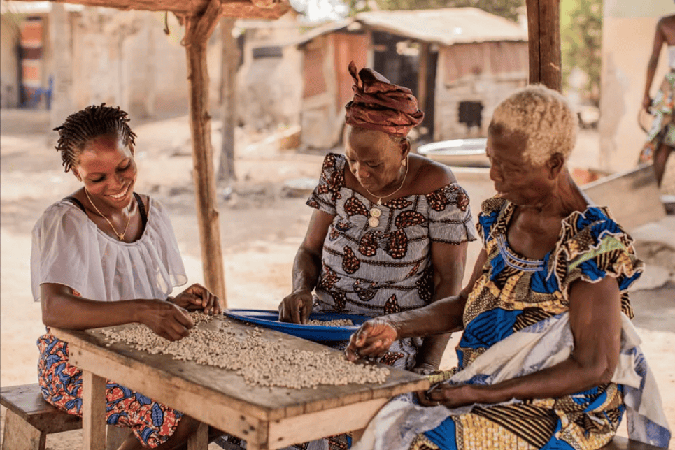 Three women from Togo — across three generations — sit at a table outdoors, shelling beans together.