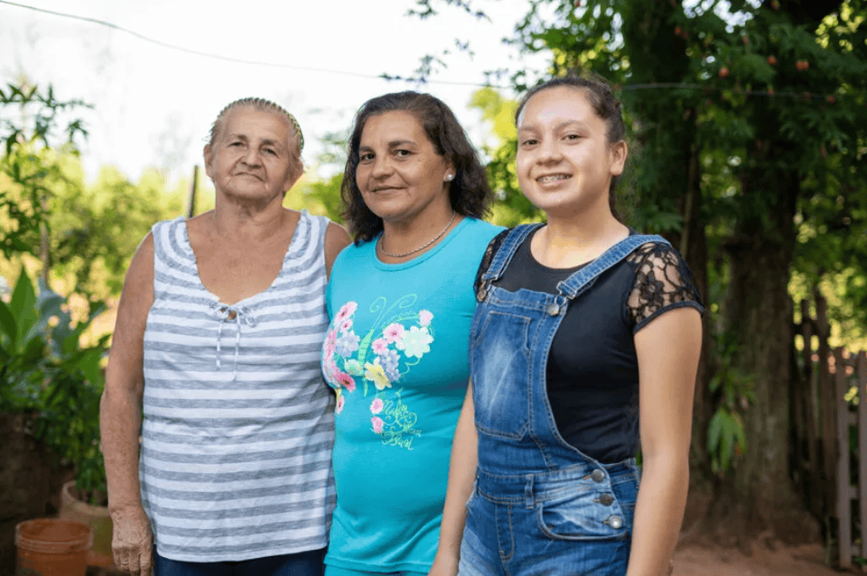 Grandmother, mother and teenage girl from Paraguay stand together outside, smiling in a rural garden setting.