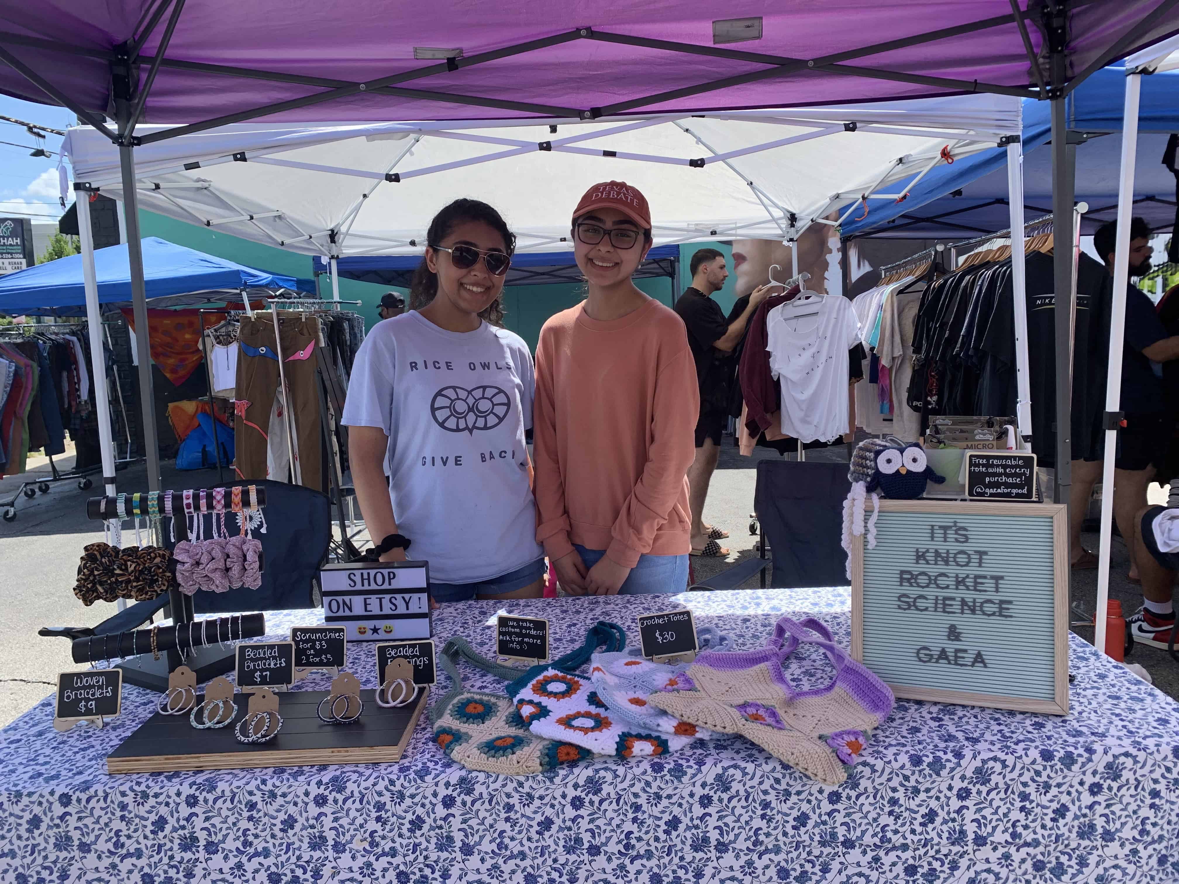 Soha stands beside a friend behind a craft booth at a local outdoor market. The table displays handmade items like scrunchies, beaded bracelets, and crocheted bags under a purple canopy. A sign on the table reads “It’s Knot Rocket Science – G.A.E.A.,” promoting her Leadership Project on environmental advocacy.