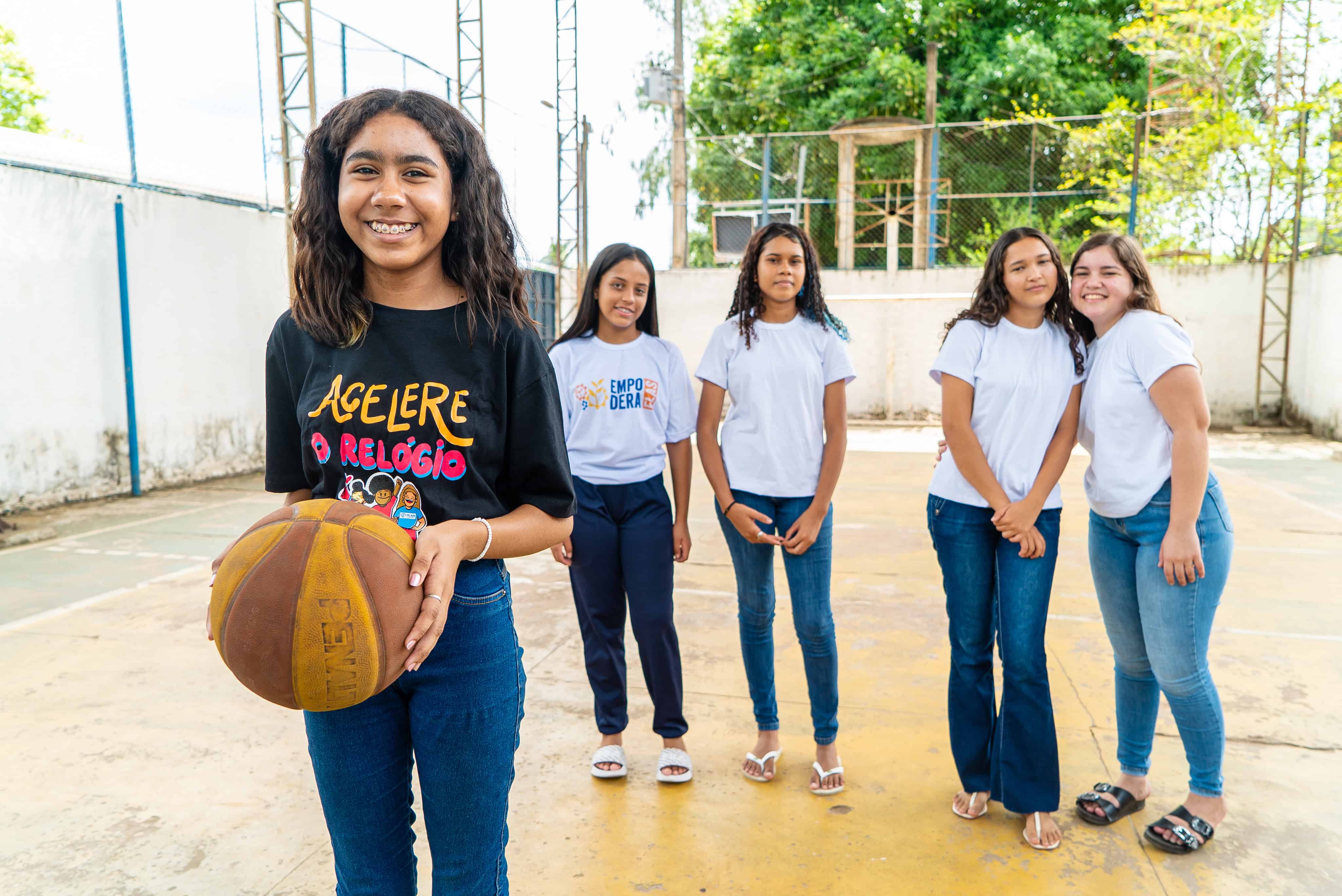 A group of teenage girls in Brazil, part of Plan’s Leadership School for Girls, stand smiling on a basketball court after a team-building activity that supports leadership and advocacy.