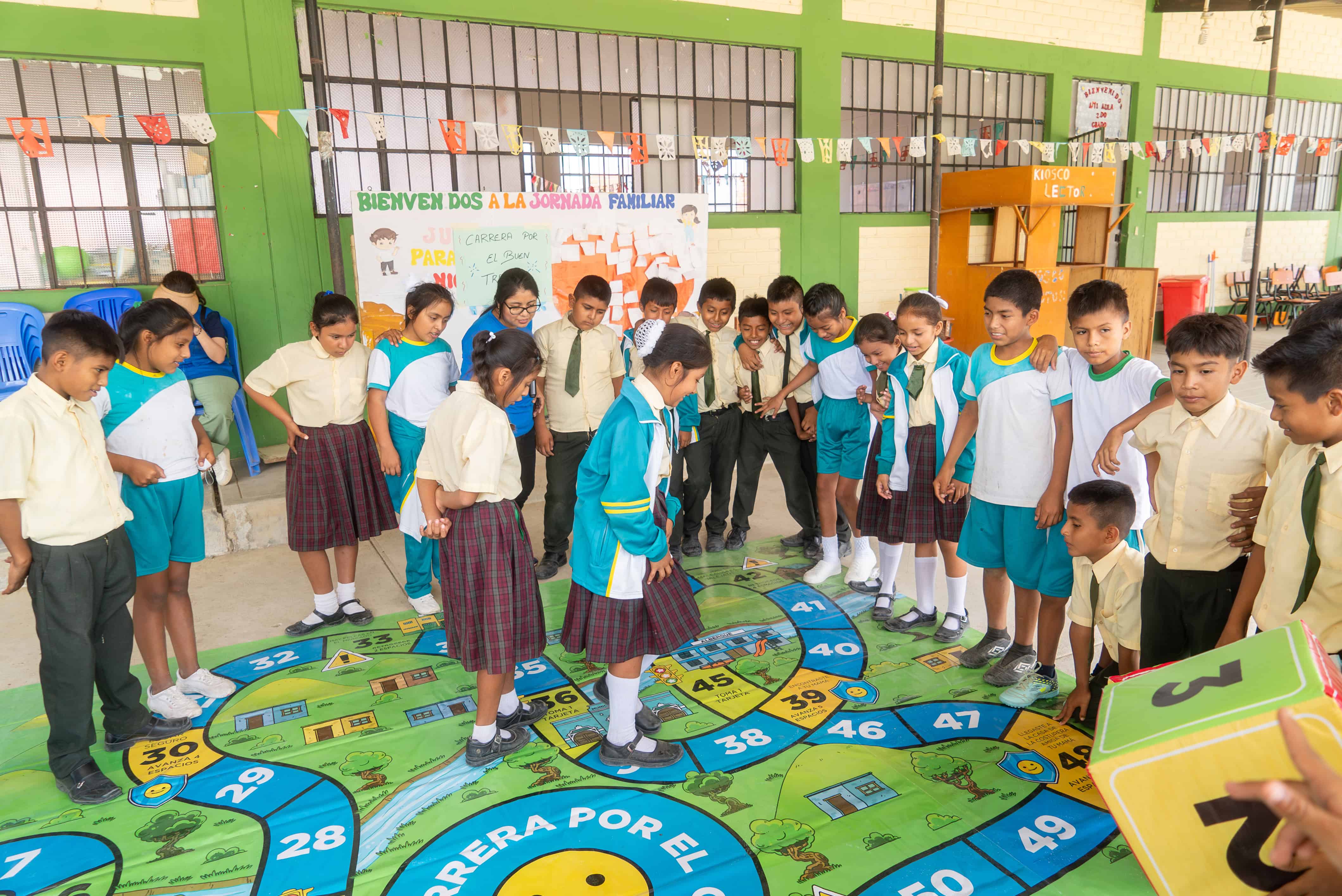 Children in school uniforms play a large floor board game during a Plan “Plan of Joy” session in Piura, Peru, designed to support emotional recovery and resilience after flooding.
