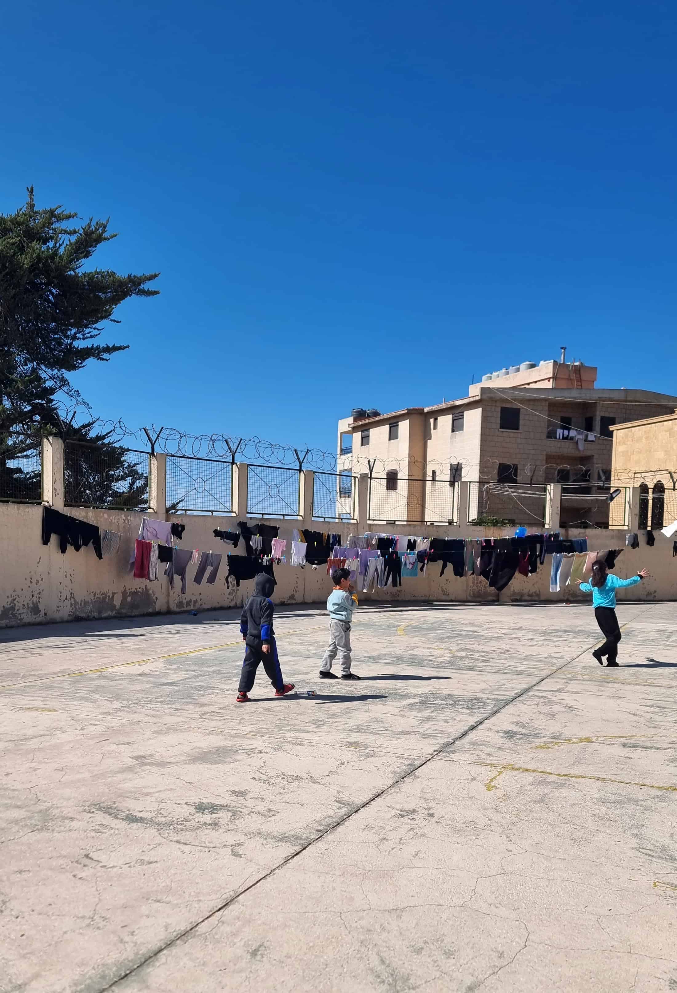 Children play in a concrete courtyard surrounded by barbed wire in Lebanon, where public schools are closed and many serve as shelters due to ongoing conflict and displacement.