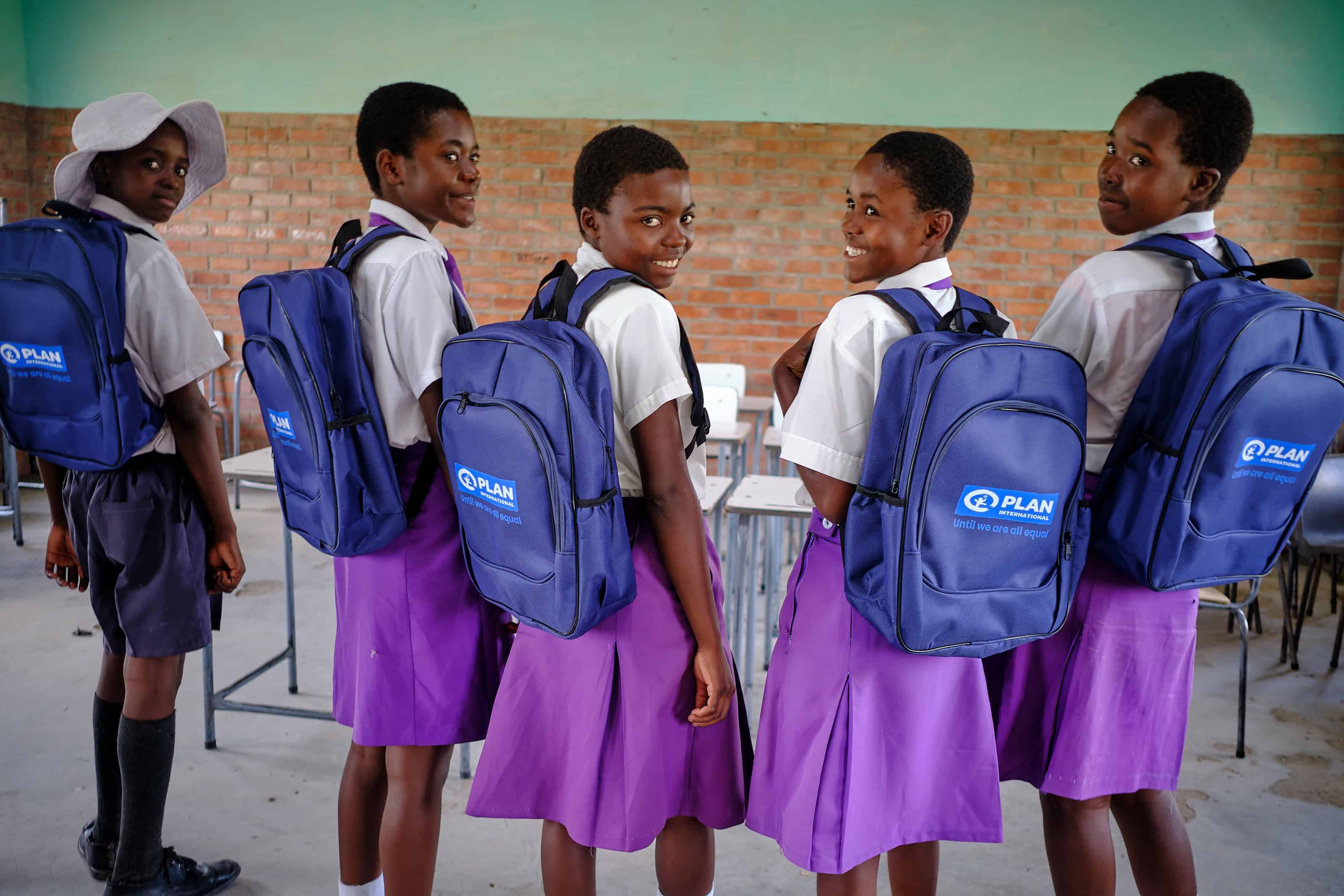 Four girls and a boy wearing school uniforms stand in a classroom facing the camera. All of the students are wearing blue Plan backpacks.