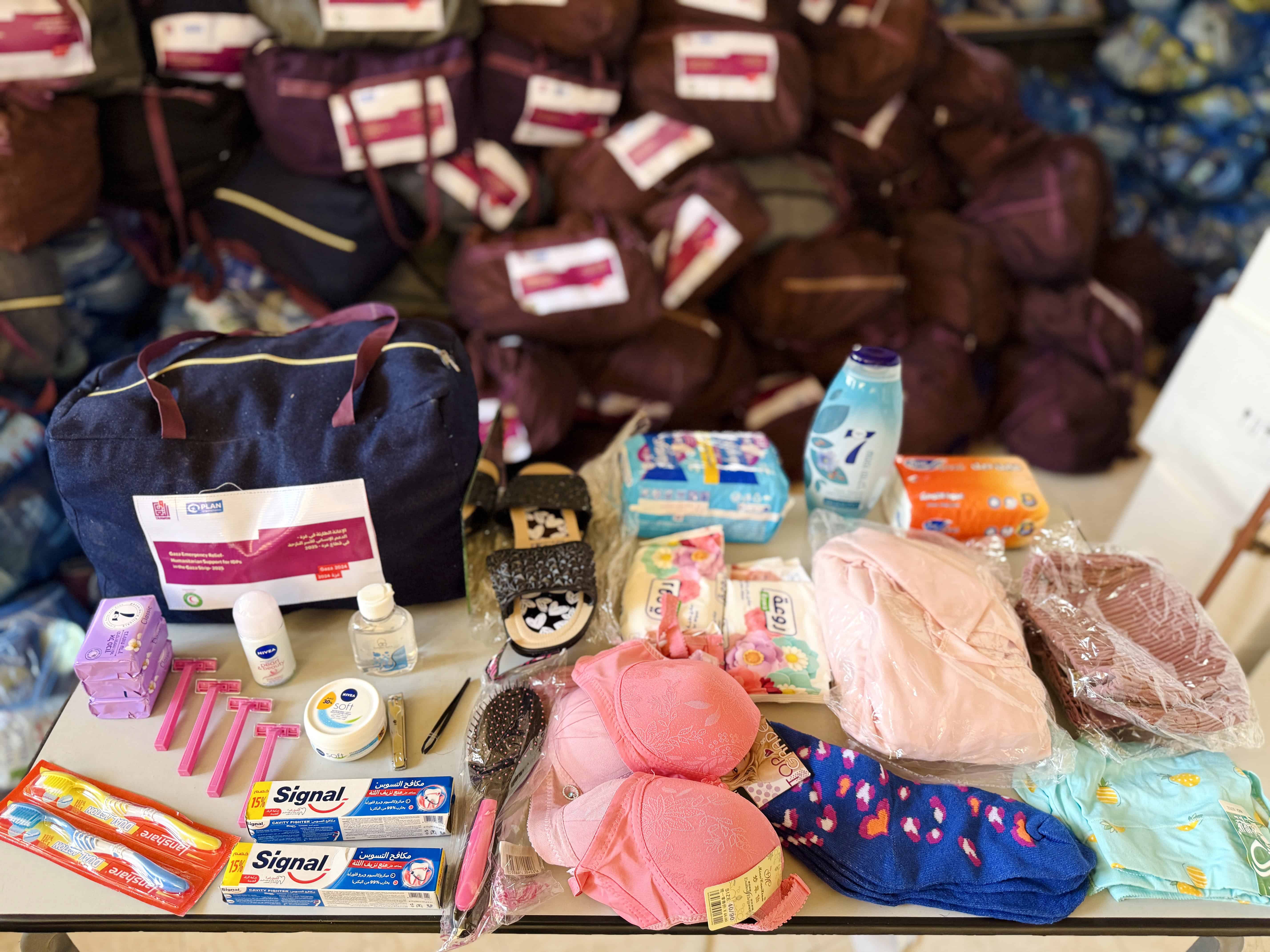 A table displays hygiene kit items in Gaza, including soap, sanitary pads, toothpaste, slippers, clothing and other supplies, with packed kits stacked behind.
