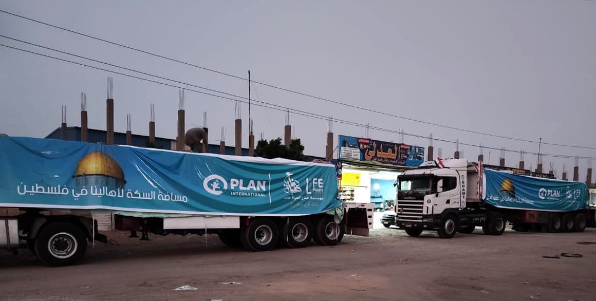 Two large trucks in Gaza covered with Plan International banners deliver food baskets to families as part of emergency humanitarian aid.
