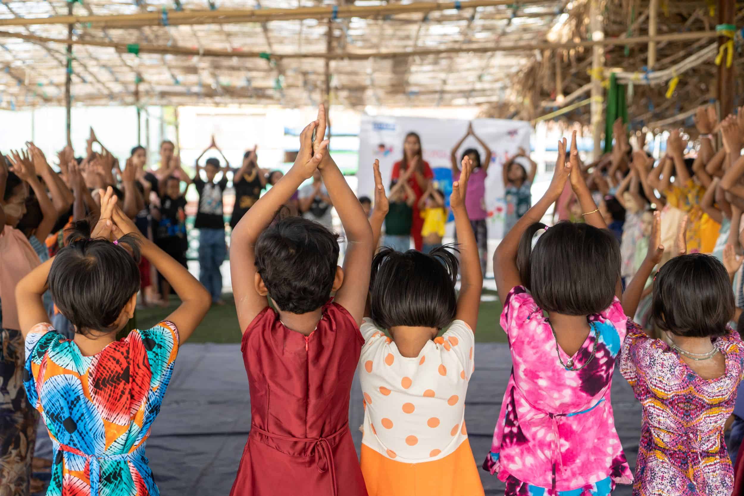 Children stand in rows inside an open shelter, raising their arms in a group activity at a child-friendly space in Mandalay, Myanmar.