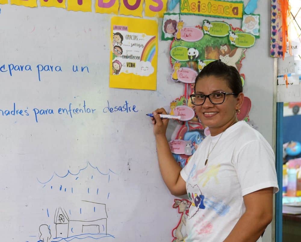A woman wearing a white t-shirt and glasses smiles as she writes on a whiteboard with a marker.