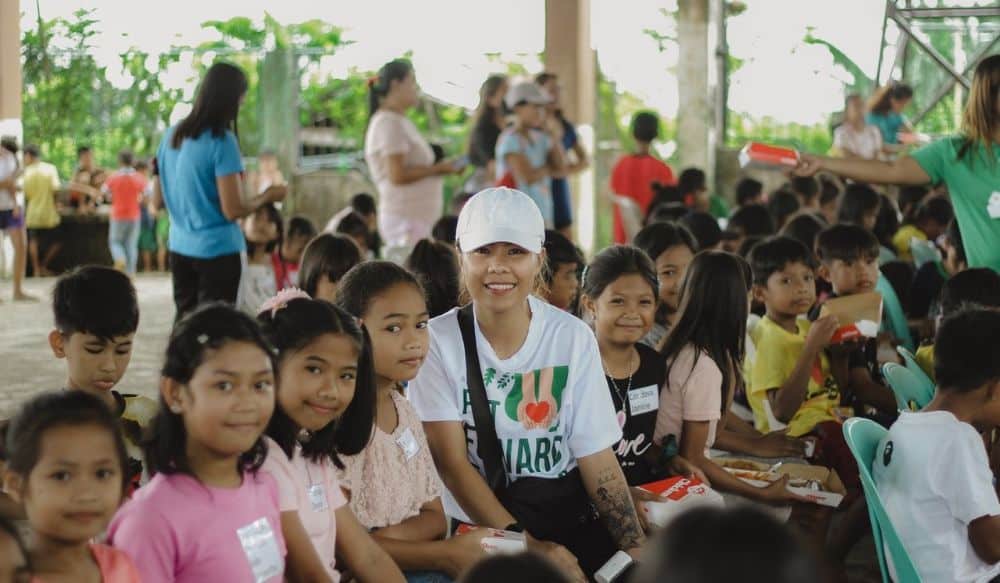 A group of children and a woman in a white hat smile together at a community event.