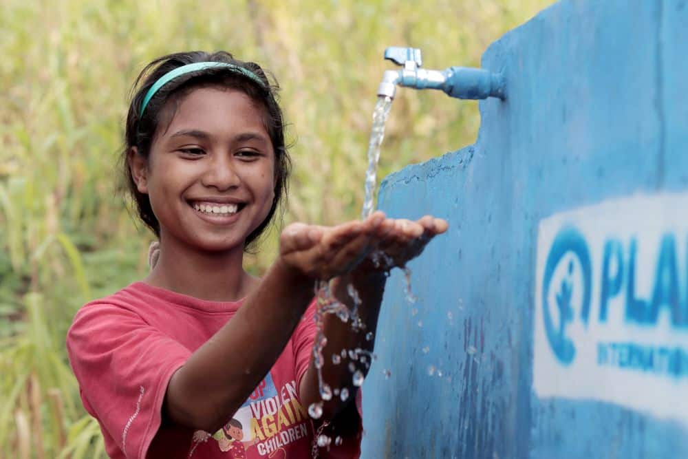 A girl smiles and holds out her hands as water flows from a tap.