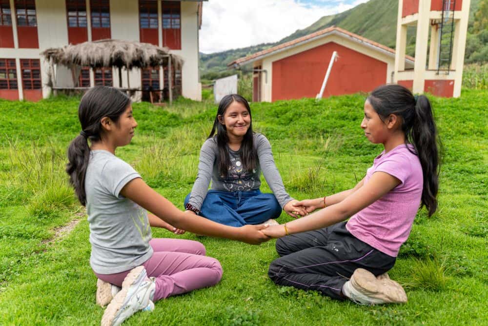 Three girls sit on the grass in a circle and hold hands.