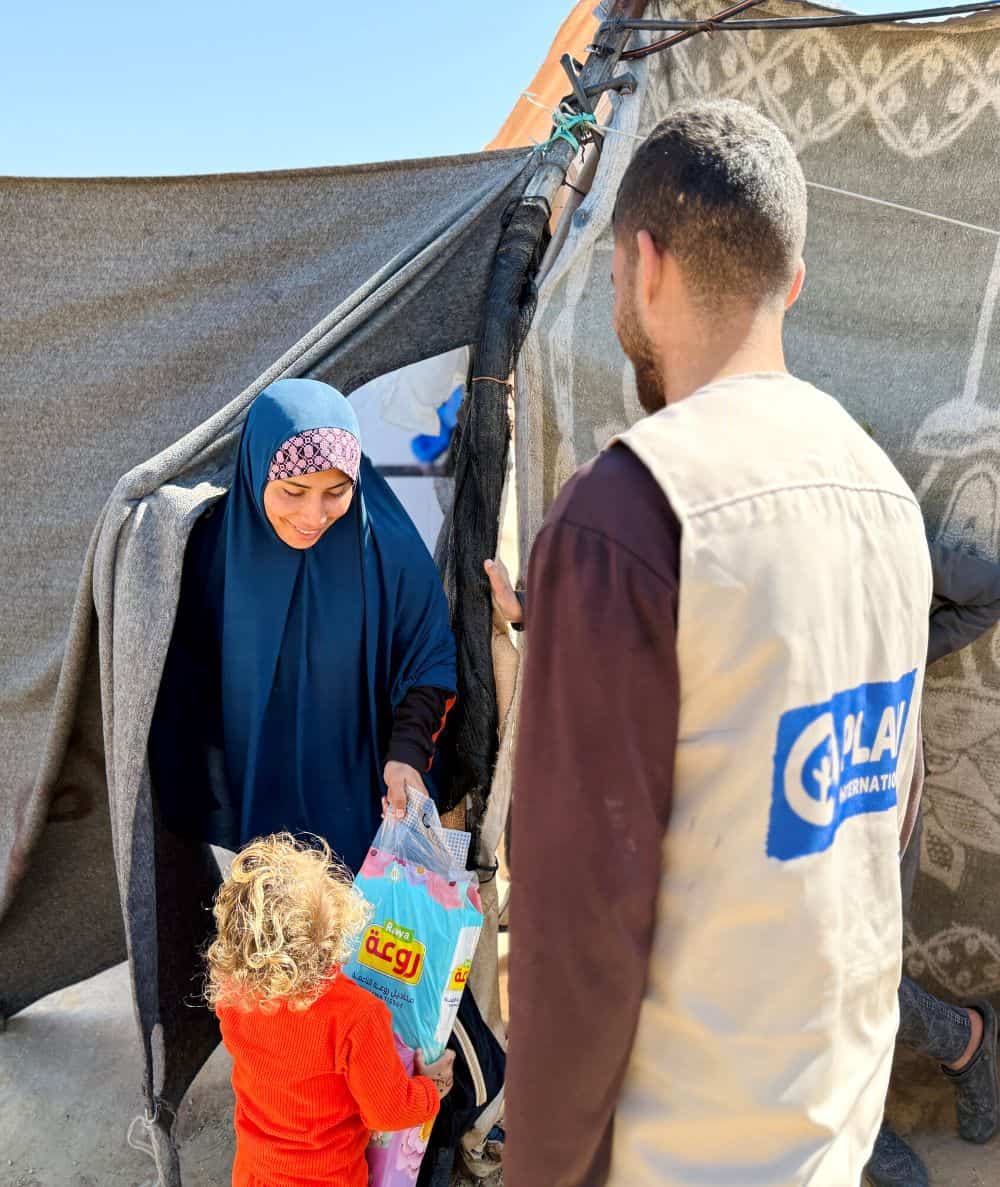 A mother stands outside at a displacement camp and smiles at her child who is holding a kit from Plan. A man wearing a vest with the Plan logo on the back stands beside them.