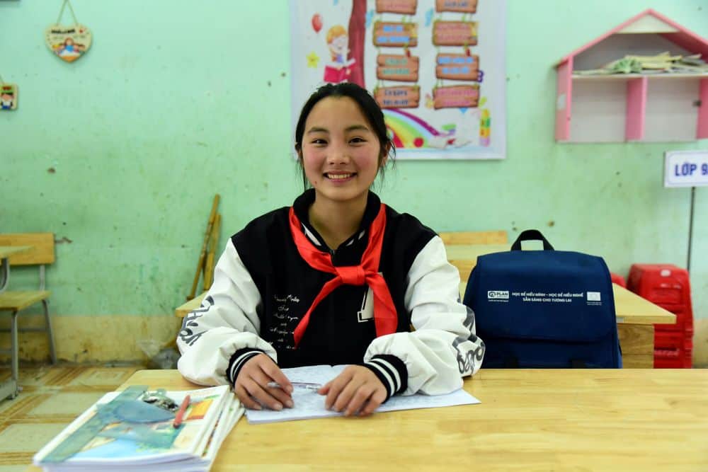 A girl smiles at her desk with a notebook and other school supplies in front of her and her backpack beside her.