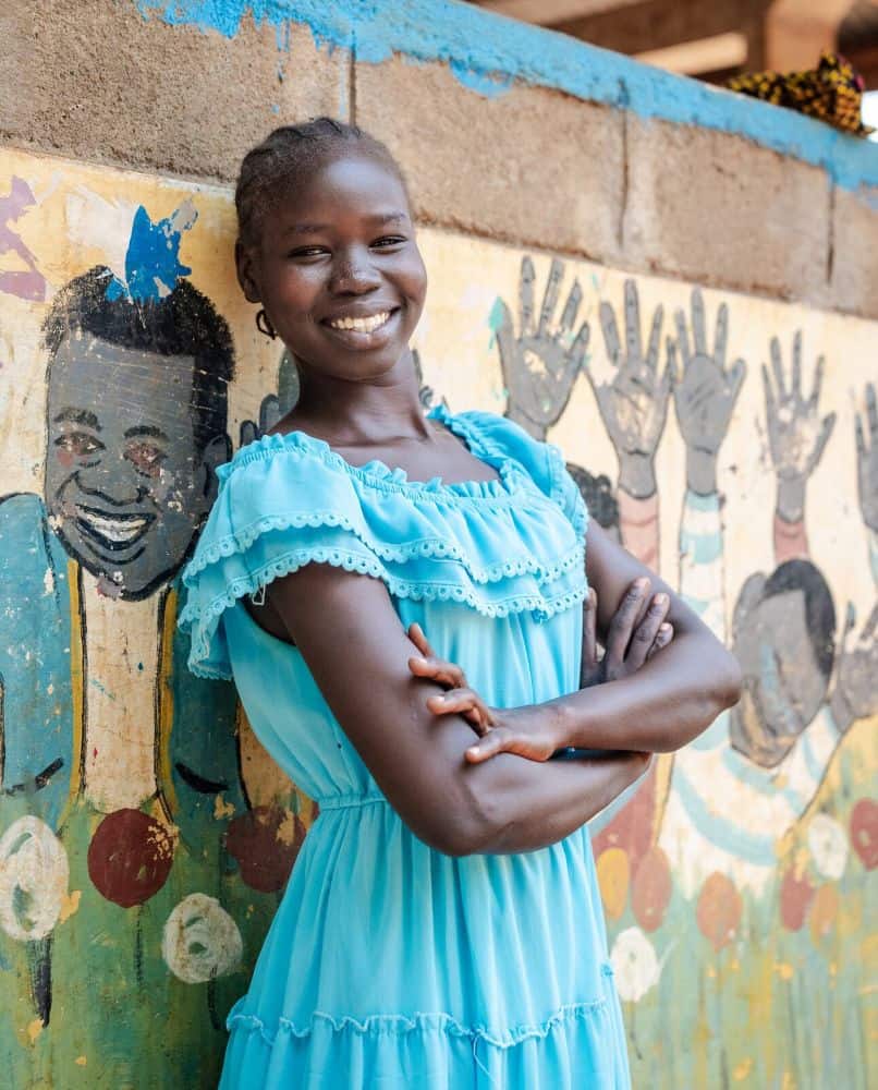 A girl wearing a blue dress leans back against a brick wall with a mural painted on it and smiles.